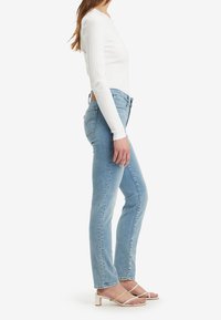 Světle modré džínové kalhoty s úzkým střihem, vysokým pasem a jemným blednutím, doplněné dlouhým rukávem bílého kabátu a bílé sandály na řemínky.