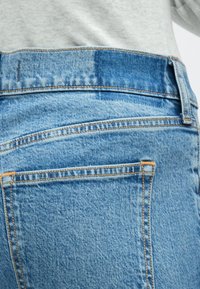 Svetlo modre kavbojke iz jeansa z klasičnim dizajnom s petimi žepi, z oranžnim šivom in rahlo izpraznjeno teksturo pasu.