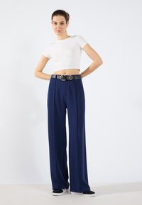 Hvit ribbet crop-top kombinert med lange, marineblå vide bukser med et svart belte med metallakenter og casual svarte joggesko.