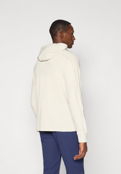 Krémová mikina s kapucňou a textúrovanou tkaninou, vo voľnom strihu a s rebrovanými manžetami. Nosená s námorníckymi nohavicami, zobrazuje jednoduchý a funkčný dizajn.