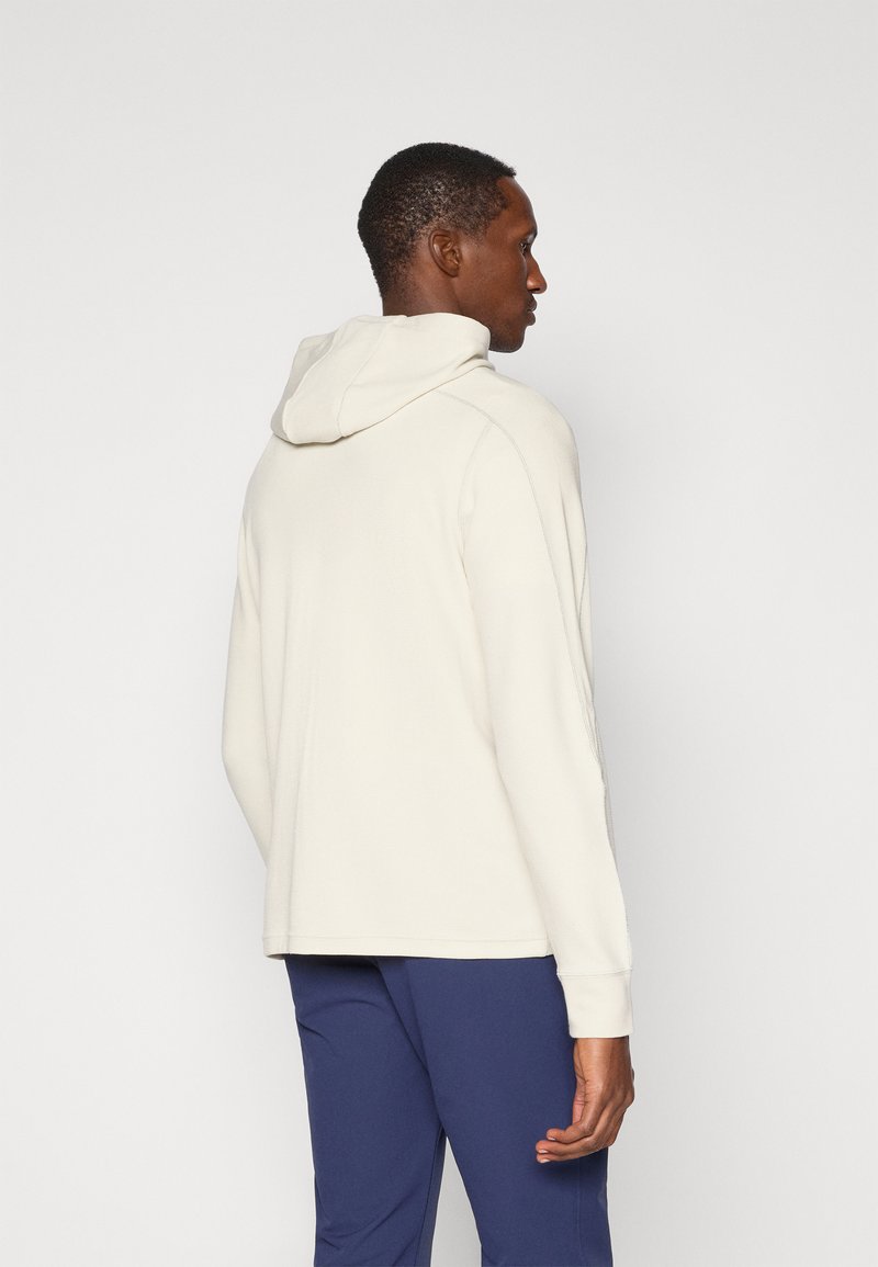 Krämfärgad hoodies med en texturerad tygkvalitet, avslappnad passform och ribbade muddar. Bärs med marinblå byxor, vilket visar en enkel och funktionell design.