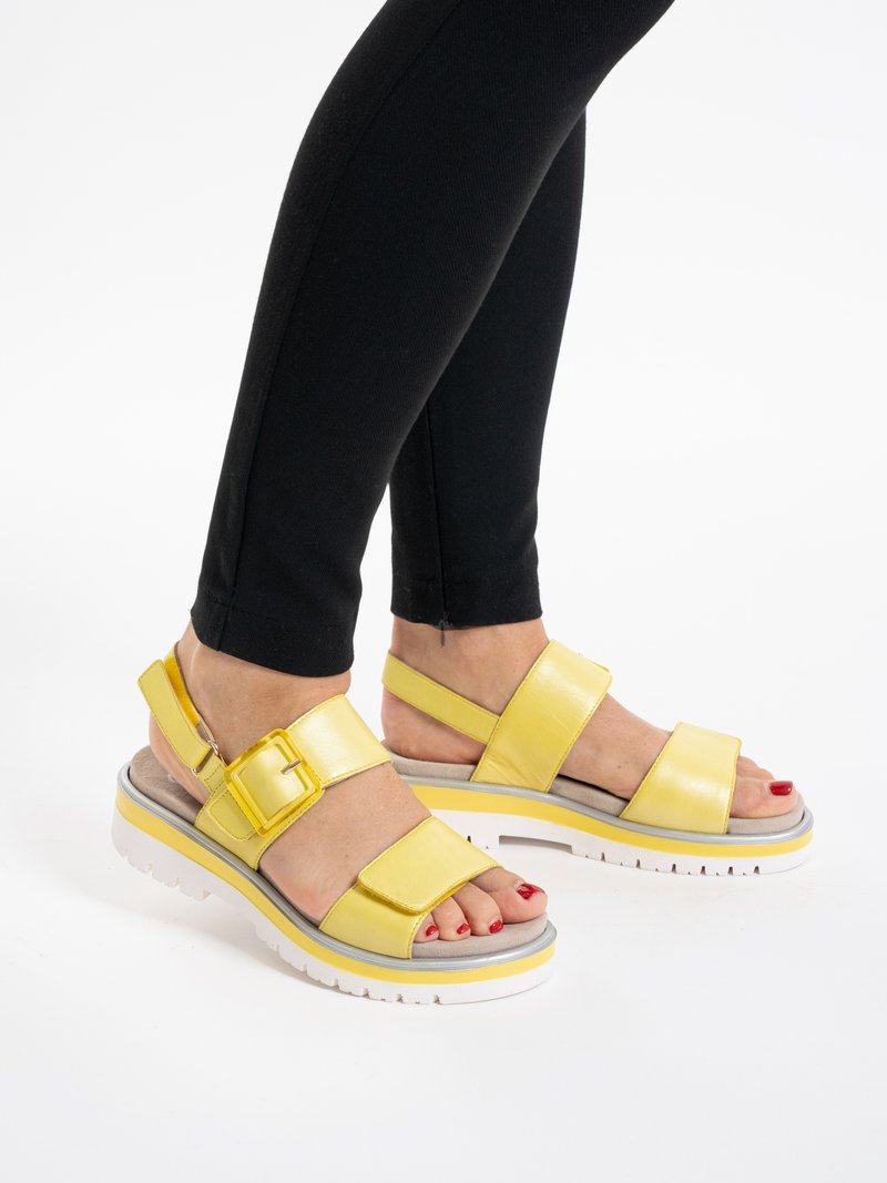 Feet wearing yellow buckle sandals with white soles and red nail polish, paired with black fitted pants.