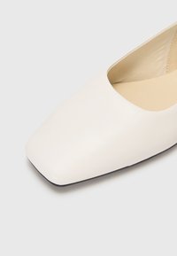 Pantofii plata din piele alba, cu varf patrat, au o textura neteda. Interiorul este captusit cu un fleece bej. Talpa este neagra, oferind un contrast cu partea superioara.