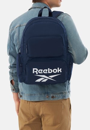 Mężczyzna w jasnoniebieskiej kurtce dżinsowej i brązowych spodniach niesie granatowy plecak Reebok z wieloma zapinanymi na zamek kieszeniami.