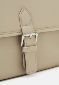 Nahaufnahme einer beigefarbenen Lederhandtasche mit einem Metallschnallenverschluss, auffälligen Nähdetails und einer glatten, strukturierten Oberfläche.