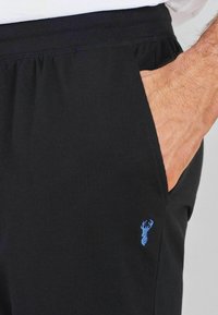 Pantaloni jogger negri dintr-un material moale, cu un elastic în talie. Au o buzunar lateral și un mic emblema al unei căprioare albastre pe partea stângă.