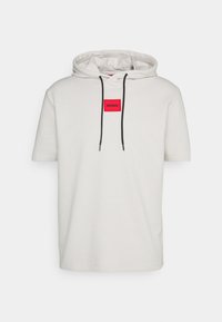 Pullover hoodie crema con maniche corte, caratterizzato da una toppa rettangolare rossa con il logo "HUGO" e lacci neri sul cappuccio.