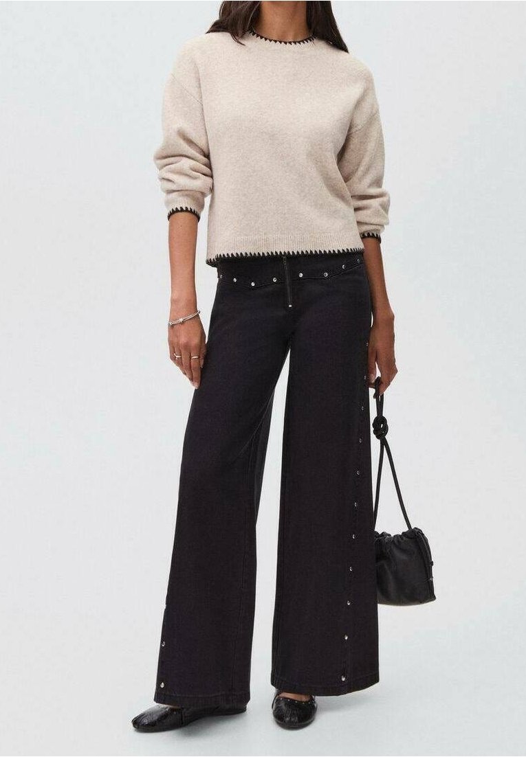 Femme portant un pull crème avec bordure noire, un pantalon noir à jambes larges avec des boutons sur les côtés, des ballerines noires et tenant un petit sac noir.