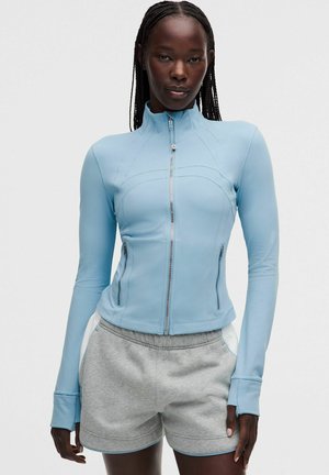 Mujer con trenzas, vistiendo una chaqueta azul claro con cremallera y pantalones cortos deportivos grises, de pie frente a un fondo claro y liso.