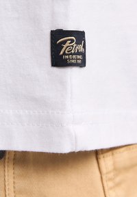 Hvid bomulds t-shirt med et sort og guld stofmærke, hvor der står "Petrol Industries Since 1983" syet i den nederste søm.