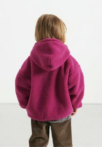 Fleece hoodie i lys pink, med en stor hætte og oversized pasform. Brune corduroy bukser bæres under. Baggrunden er ensfarvet hvid.