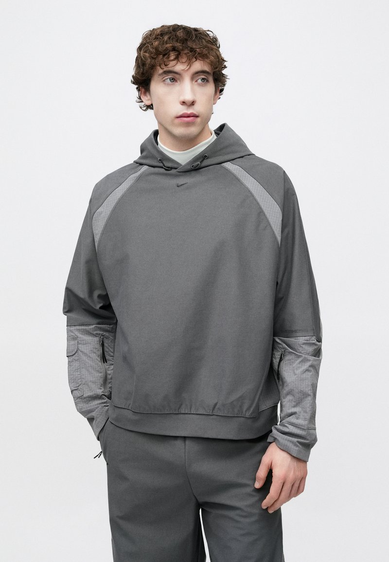 Grå pullover-hoodie med en frontlomme, snørelukning på hætten og teksturerede detaljer på ærmerne. Glat stof med en afslappet pasform og geometriske mønstre.