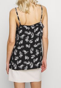 Blond kvinna i en svart ärmlös topp med vita blommönster och en ljusbeige kjol, sedd bakifrån mot en enfärgad bakgrund.