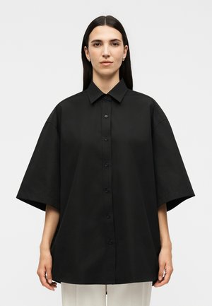 Γυναίκα με ίσια σκούρα μαλλιά, που φοράει υπερμεγέθη μαύρο πουκάμισο με κοντά μανίκια και λευκό παντελόνι, στέκεται μπροστά σε απλό φωτεινό φόντο.