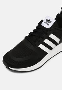 Svart idrottssko med mesh-övre, tre vita ränder på sidan och gummisula; har snörning och vadderad krage.