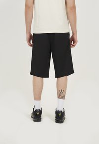 Black athletic shorts with a relaxed fit, reaching mid-calf, paired with black trainers and white ankle socks, showing a tattoo on the leg.