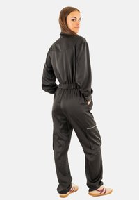 Combinaison noire ajustée avec taille élastique, manches longues et poches zippées ; texture lisse ; portée avec des chaussures de couleur claire.