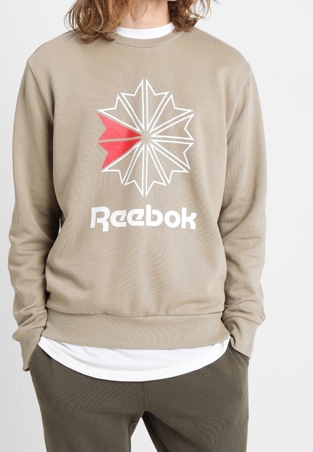 Personne portant un sweat-shirt beige Reebok avec un logo étoile blanc et rouge, un sous-vêtement blanc et un pantalon vert foncé, les mains dans les poches.