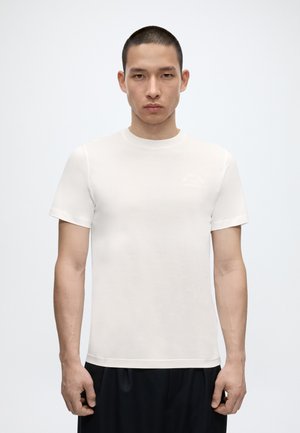 Άνδρας με κοντά μαλλιά που φοράει απλό λευκό κοντομάνικο μπλουζάκι και μαύρο παντελόνι, στέκεται μπροστά σε λευκό φόντο.