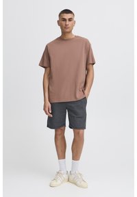 Maglietta marrone a maniche corte in cotone, abbinata a pantaloncini grigio scuro. Il modello indossa calzini bianchi e sneakers color crema. Design minimalista.