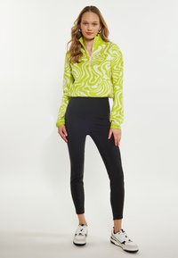 Grüner und weißer, gemusterter Fleece-Pullover mit einem halben Reißverschluss-Kragen, kombiniert mit schwarzen, schmal geschnittenen Hosen und weißen Sneakers mit schwarzen Akzenten.
