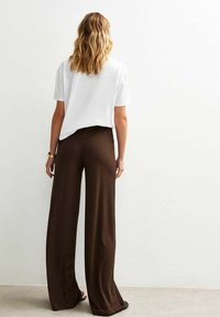 Een vrouw met golvend haar draagt een witte t-shirt en bruine geplooide broek, staande op een lichtgekleurde ondergrond tegen een neutrale achtergrond.
