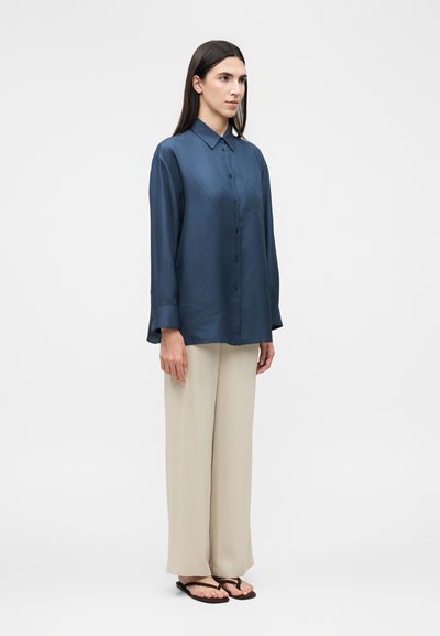 Γυναίκα με μπλε σκούρο πουκάμισο με μακριά μανίκια και κουμπιά, μπεζ παντελόνι με φαρδιά πόδια και μαύρα σανδάλια, στέκεται μπροστά από έναν απλό ανοιχτόχρωμο φόντο.