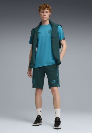 Junger Mann, der tealfarbene Manchester City Sportbekleidung trägt, darunter ein T-Shirt, Shorts, weiße Socken, schwarze Puma-Sneaker und einen Pullover über den Schultern.
