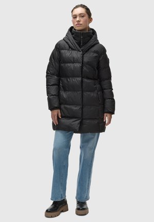 Zwarte pufferjas met een capuchon, voorzien van een gequilt design, een rits aan de voorkant en zijzakken. Gecombineerd met lichtblauwe jeans en zwarte schoenen.