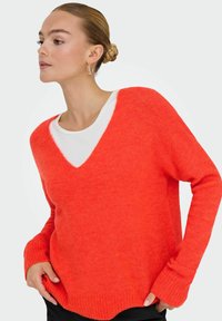 Femme avec chignon tressé blond portant des boucles d'oreilles créoles dorées, un pull col V orange vif sur une chemise blanche, mains près des poches.