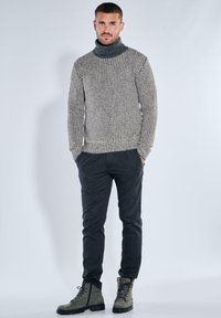 Strickpullover mit Stehkragen in Hellgrau und einem dunkleren strukturierten Muster, kombiniert mit dunklen Slim-Fit-Hosen und grauen Schnürboots.