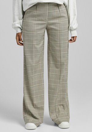 Personne portant un pantalon à carreaux taille haute, larges, dans des teintes beige, noir et blanc, associé à des baskets blanches et un sweat-shirt gris clair.