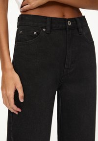 Gros plan sur une personne portant un jean en denim noir taille haute et un haut blanc court, main reposant à côté de la hanche.