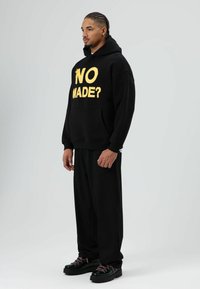 Zwarte hoodie met gele tekst "NO MADE?" aan de voorkant, gecombineerd met zwarte joggers en zwarte schoenen met roze veters. Soepele stof, casual pasvorm.