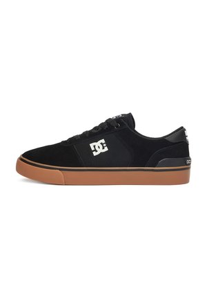 Sneaker nero basso con suola in gomma marrone, logo DC bianco sul lato, lacci neri e collarino imbottito sul tallone.