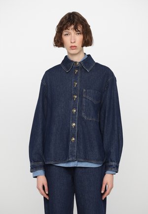 Giacca in denim blu scuro con chiusura a bottoni, colletto aperto e taschino sul petto. Presenta maniche ampie e un orlo leggermente accorciato.