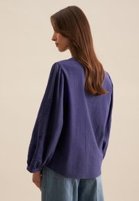 Blusa a maniche lunghe viola realizzata in tessuto leggero. Presenta dettagli ricamati sulle maniche e una vestibilità comoda.