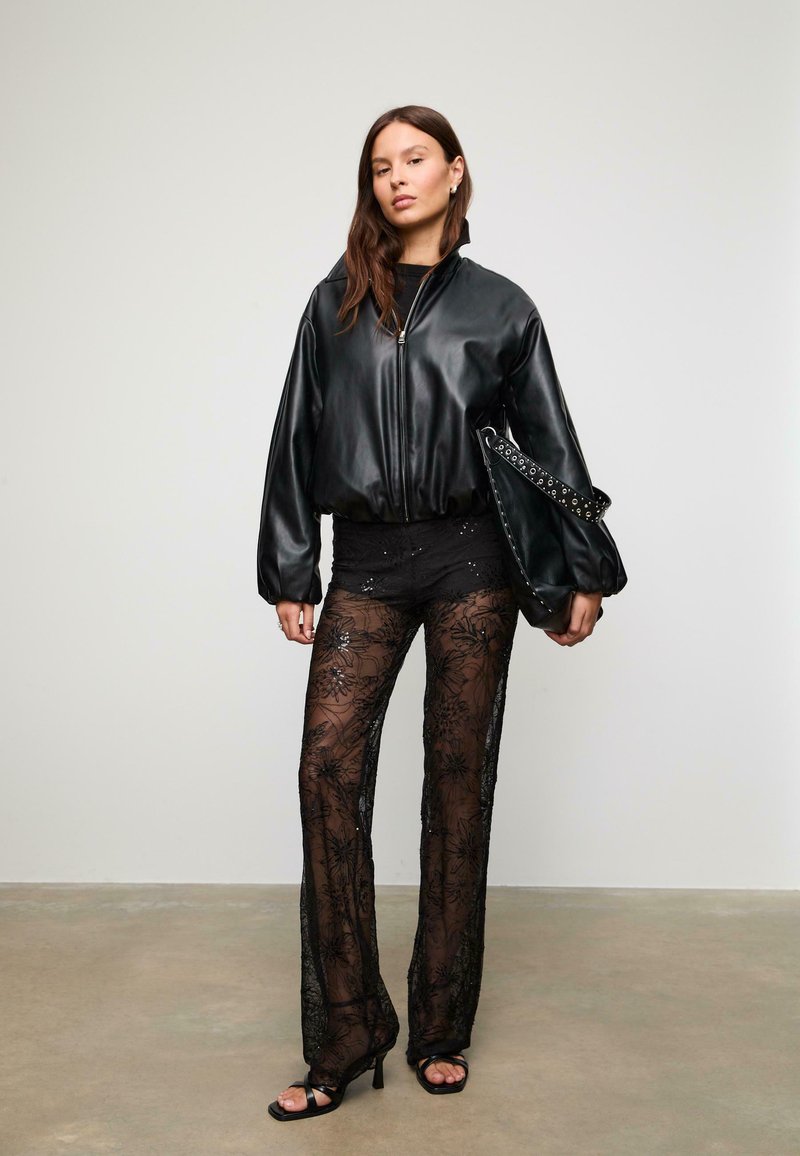 Zwarte, nep leren bomberjack met een cropped silhouet; doorschijnende zwarte kanten broek met bloemmotieven; zwarte gespenhakken; bijpassende tas.