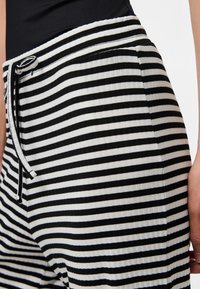 Musta-valkoiset vaakaraitaiset housut, joissa on kiristysnyöri vyötäröllä, yhdistetty mustaan yläosaan. Kuvassa oikea lantio ja käsi.