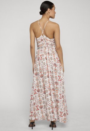 Frau in einem langen weißen Blumenkleid mit dünnen gekreuzten Trägern und schwarzen Sandalen mit hohen Absätzen, stehend vor einem schlichten Hintergrund.