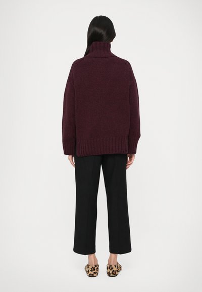 Femme se tenant de dos, portant un pull à col roulé oversized violet foncé, un pantalon court noir et des chaussures à enfiler imprimé léopard.