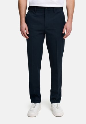 Mann trägt schwarze Slim-Fit-Hose, weiße Sneakers und ein weißes, in die Hose gestecktes T-Shirt vor schlichtem Hintergrund.