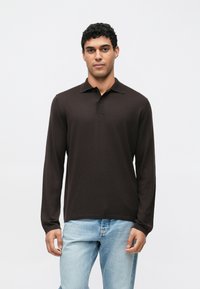 Polo a maniche lunghe in marrone scuro, realizzata in tessuto strutturato. Presenta un colletto e due bottoni sul collo. Abbinata a jeans azzurri.