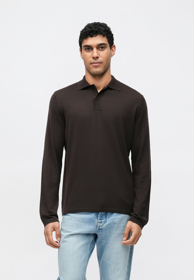 Polo a maniche lunghe in marrone scuro, realizzata in tessuto strutturato. Presenta un colletto e due bottoni sul collo. Abbinata a jeans azzurri.