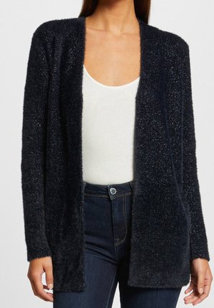 Donna che indossa un cardigan nero aperto e soffice su una canottiera bianca con scollo rotondo e jeans a vita alta blu scuro.