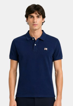 Uomo che indossa una polo blu navy a maniche corte con piccolo logo a toppa sul petto sinistro, in piedi davanti a uno sfondo chiaro e semplice.