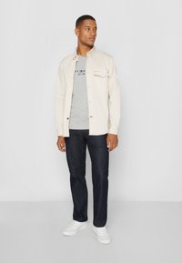 Ljus beige skjorta med knäppning över en grå t-shirt med text, mörkblå jeans och vita sneakers. Rakt snitt med minimalistisk design.