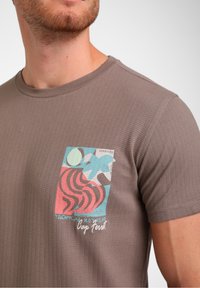 Bruine T-shirt met structuur, featuring een meerkleurig grafisch patch met abstracte patronen, waaronder zwirls en cirkels, en geborduurde tekstdetails.