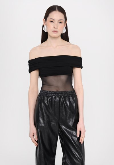 Melnā off-the-shoulder blūze ar rūcinājuma detaļām un caurspīdīgu acu paneli, apvienota ar melnām spīdošām ādas biksēm.