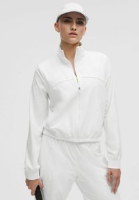 Vit zip-up jacka i ett mjukt och lätt material, med elastisk midja och krage. Har långa ärmar och en avslappnad passform.